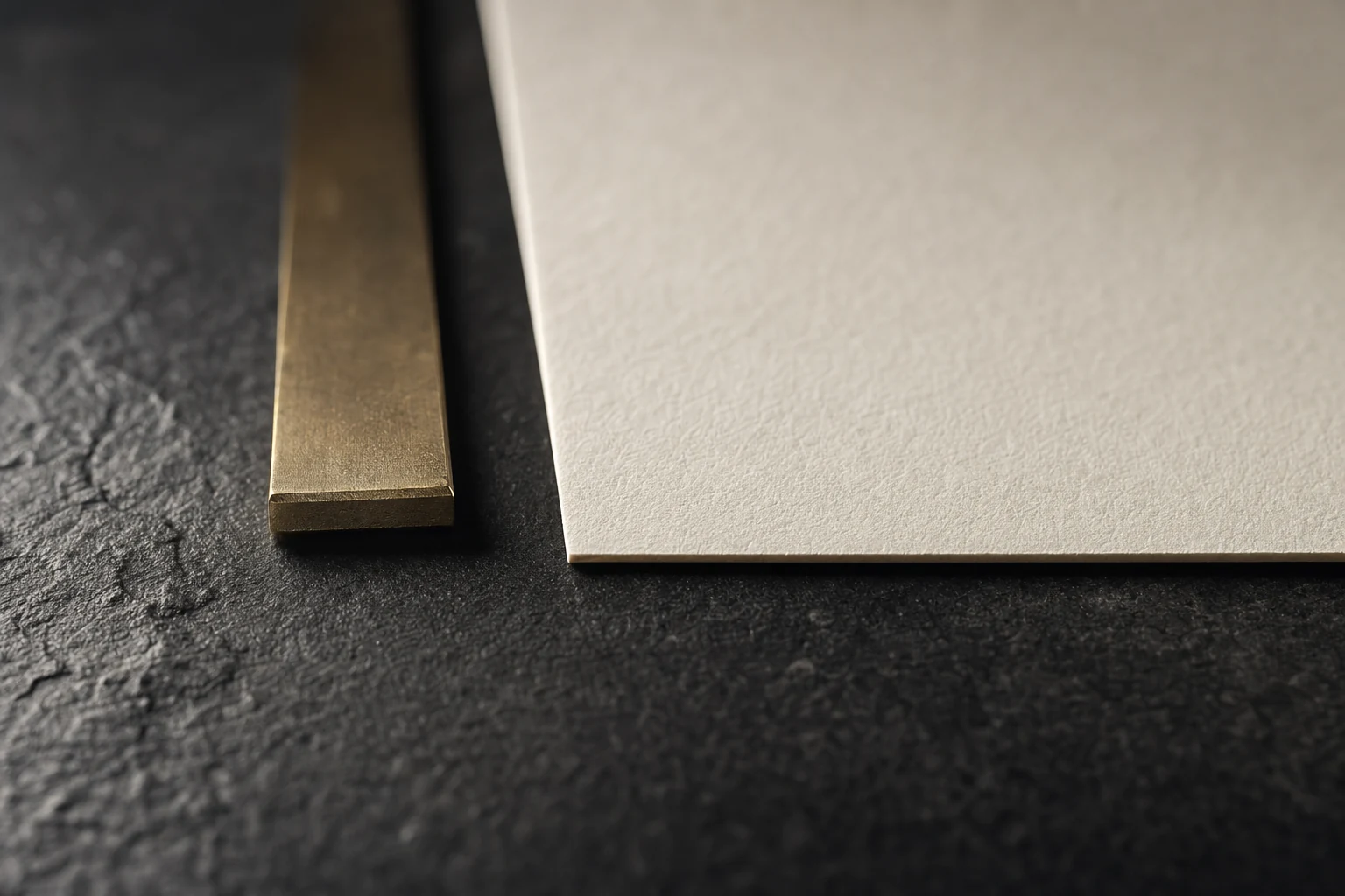 A blank sheet of heavy ivory paper beside a solid brass straightedge on black stone